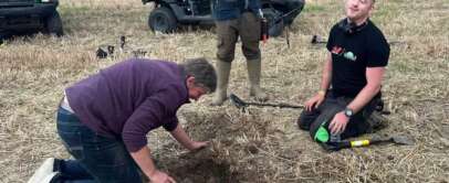 Three people in a field, one kneeling and digging in the soil while the other kneels with a metal detector, smiling. A person stands to the side. Two off-road vehicles are in the background on a grassy area.