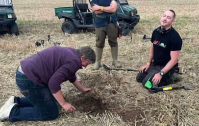 Three people in a field, one kneeling and digging in the soil while the other kneels with a metal detector, smiling. A person stands to the side. Two off-road vehicles are in the background on a grassy area.