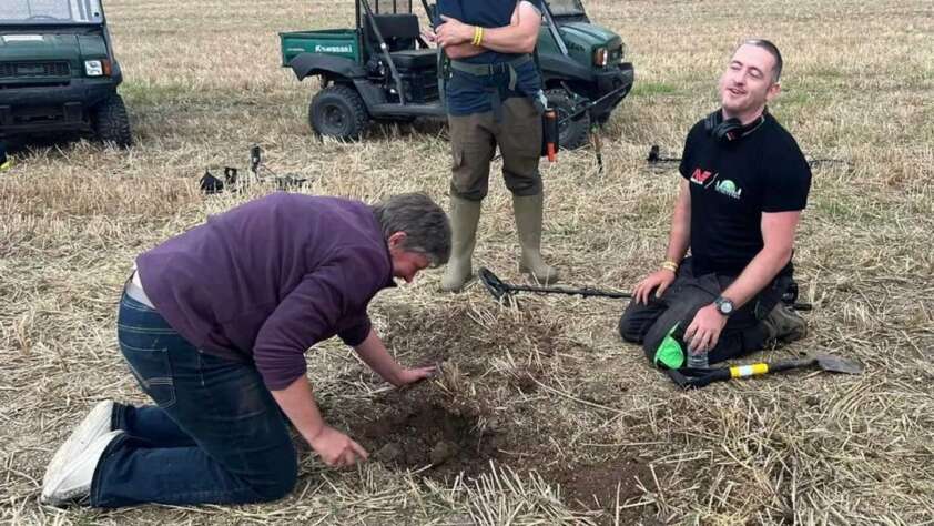 Three people in a field, one kneeling and digging in the soil while the other kneels with a metal detector, smiling. A person stands to the side. Two off-road vehicles are in the background on a grassy area.