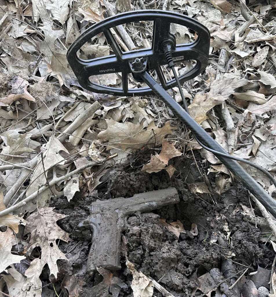 A metal detector hovers over a muddy, partially unearthed handgun on the forest floor, surrounded by dry leaves and twigs.