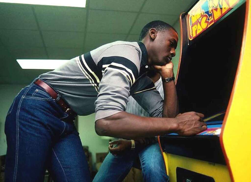 A person intensely focused, playing an arcade game. They are standing with one hand on their chin, wearing a striped sweater and jeans, in a room with a tiled ceiling. The game cabinet is yellow with a colorful display.