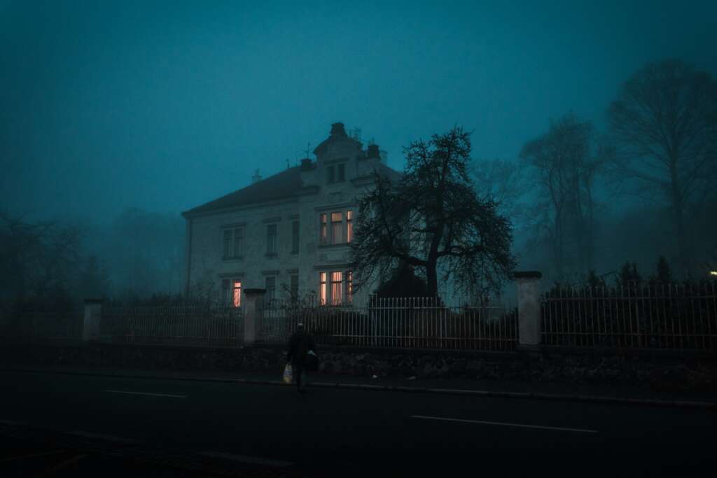 A mysterious, dimly lit house stands amid dense fog at night. Its windows emit a warm glow. Silhouetted bare trees surround it, and a lone figure walks on the dark road in front, carrying a bag.