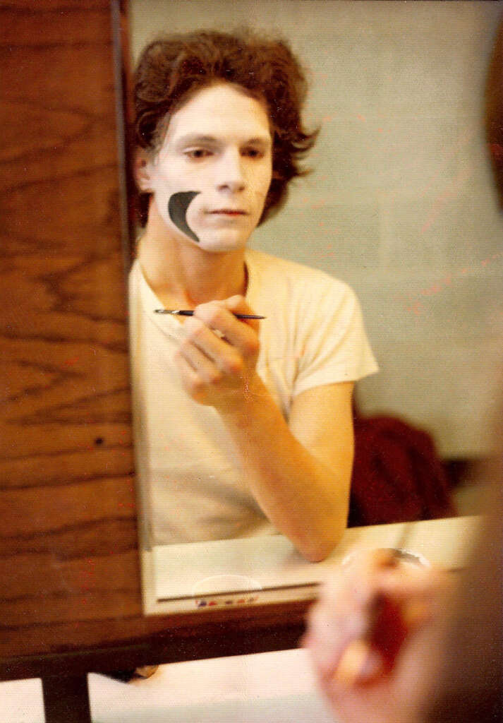 Person applying white face paint with a brush while looking in a mirror. The person is wearing a white T-shirt, and there is a black design on the right cheek. Reflections show a wooden frame and a countertop with items.