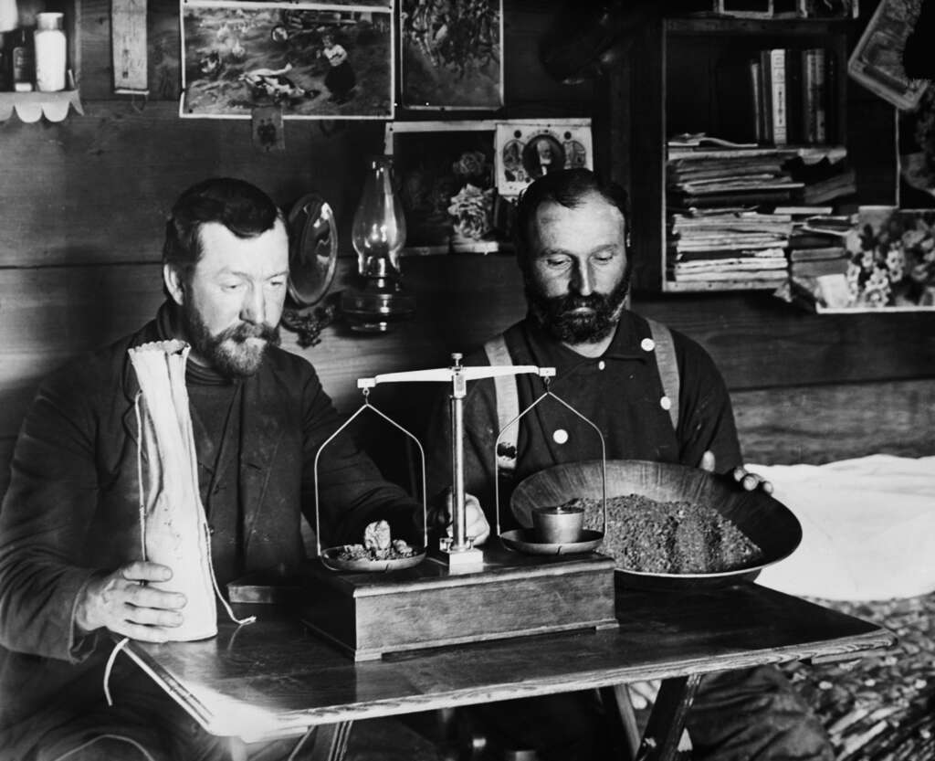 Two men in a rustic room weigh gold nuggets on a scale. One holds a bag, while the other balances a pan with gold flecks. Photos and papers hang and sit on the walls and shelves behind them.