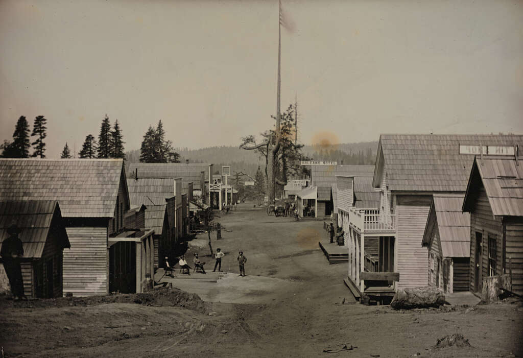 A sepia-toned historical photograph shows a dirt street lined with wooden buildings on both sides. Several people are seen walking and standing on the street. Trees are visible in the background, and a tall flagpole stands in the distance.