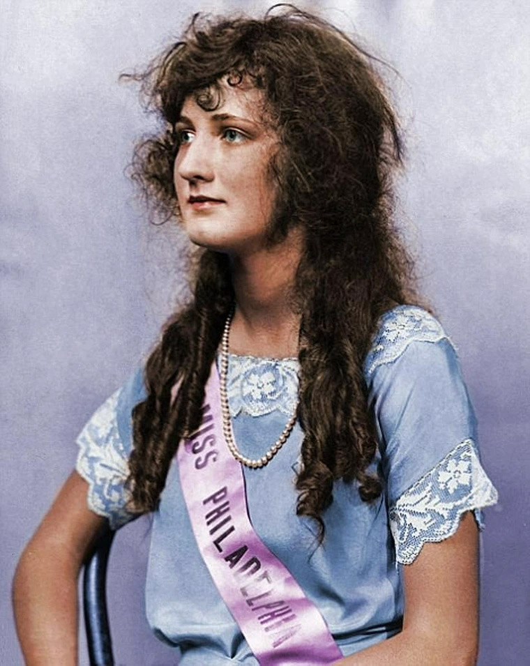 A woman with long curly hair sits on a chair, wearing a blue dress with lace sleeves and a pearl necklace. She has a sash reading "Miss Philadelphia." The background is a soft lavender color.