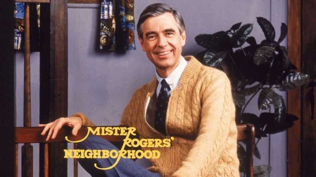 A man in a light brown cardigan and tie sits on a wooden bench in a cozy environment. Behind him, a plant and patterned curtain are visible. The text "Mister Rogers' Neighborhood" is in the foreground.