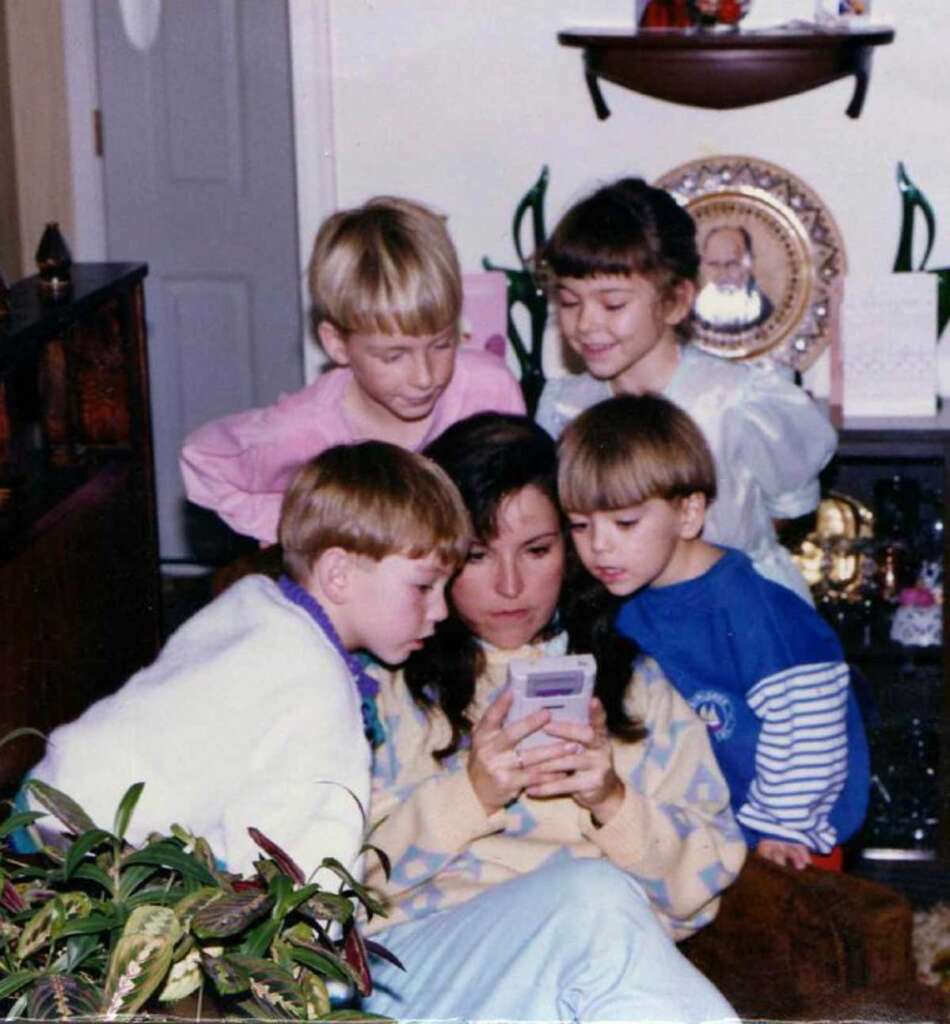 A woman is seated, holding a Game Boy, surrounded by four young children who are watching intently. One child wears a white sweater, another a blue one. There are plants and a shelf with a framed photo in the background.