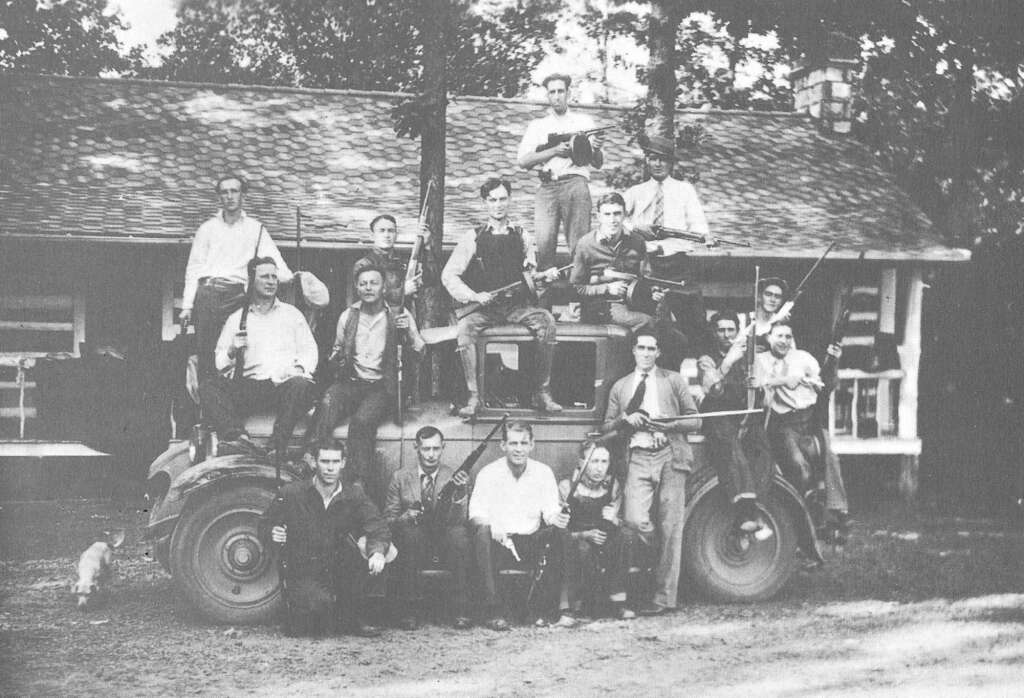 A group of sixteen people pose on and around a tractor in front of a house. They hold musical instruments, including a guitar and a fiddle. The black and white image has a vintage, rustic feel. A small dog is visible to the left.