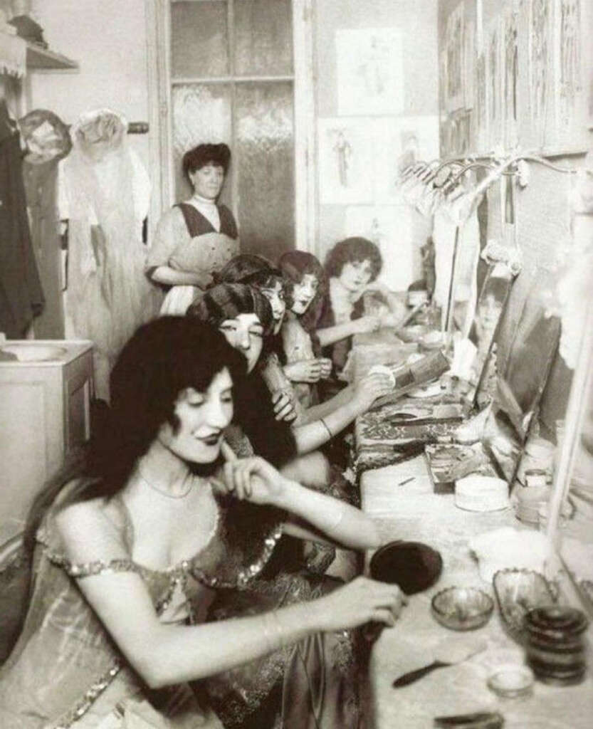 A vintage photo shows several women seated at a long dressing table, applying makeup in front of mirrors. They are dressed in ornate gowns, and a woman stands in the background. The setting appears lively, with various beauty items scattered around.