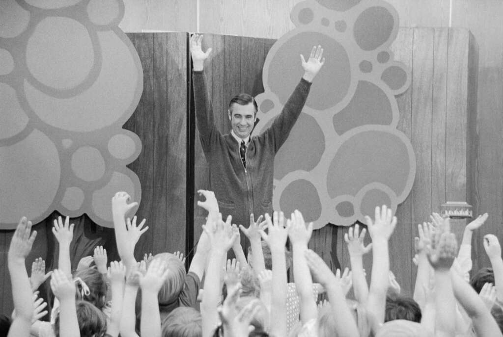 A man in a cardigan, smiling with arms raised, stands in front of a wooden background with circular patterns. In the foreground, children raise their hands, reaching toward him. The scene is lively and joyful.