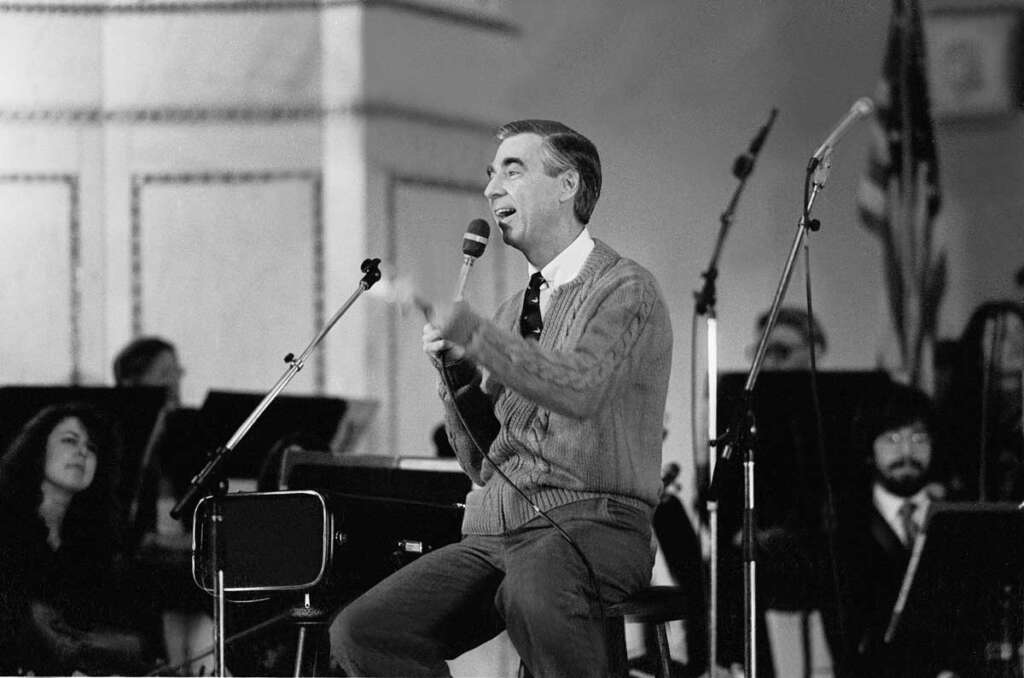 A person sitting on a stool, speaking into a microphone. They are wearing a sweater and tie. The setting appears to be a stage with an orchestra in the background. The scene is in black and white.