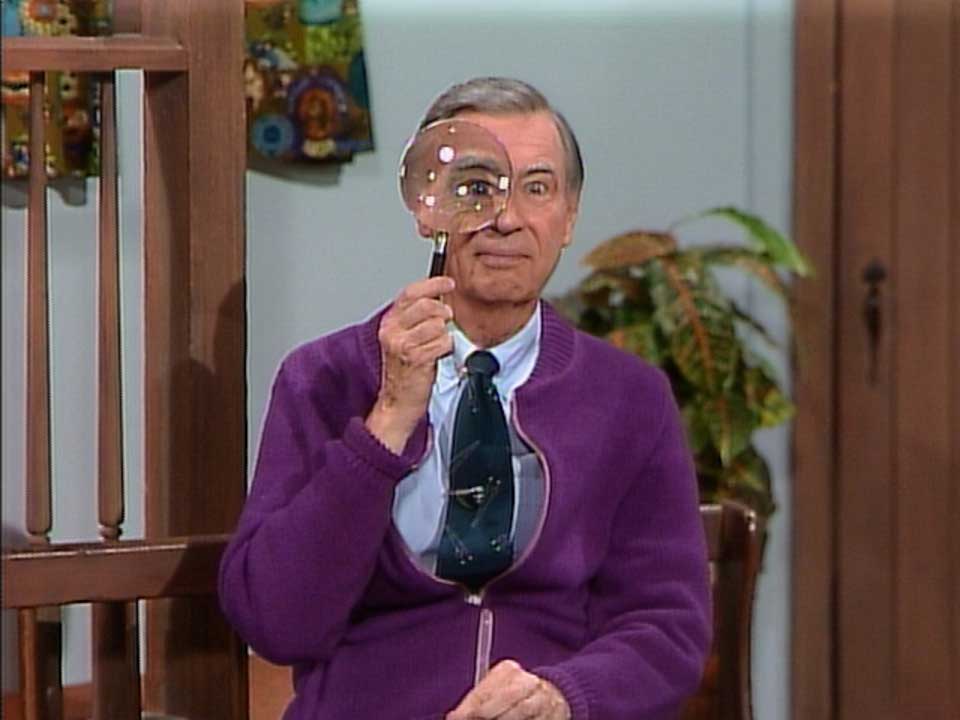 An older man sits indoors, holding a magnifying glass to his face, which distorts his eye. He wears a purple cardigan over a green tie, and there is a decorative plant in the background.