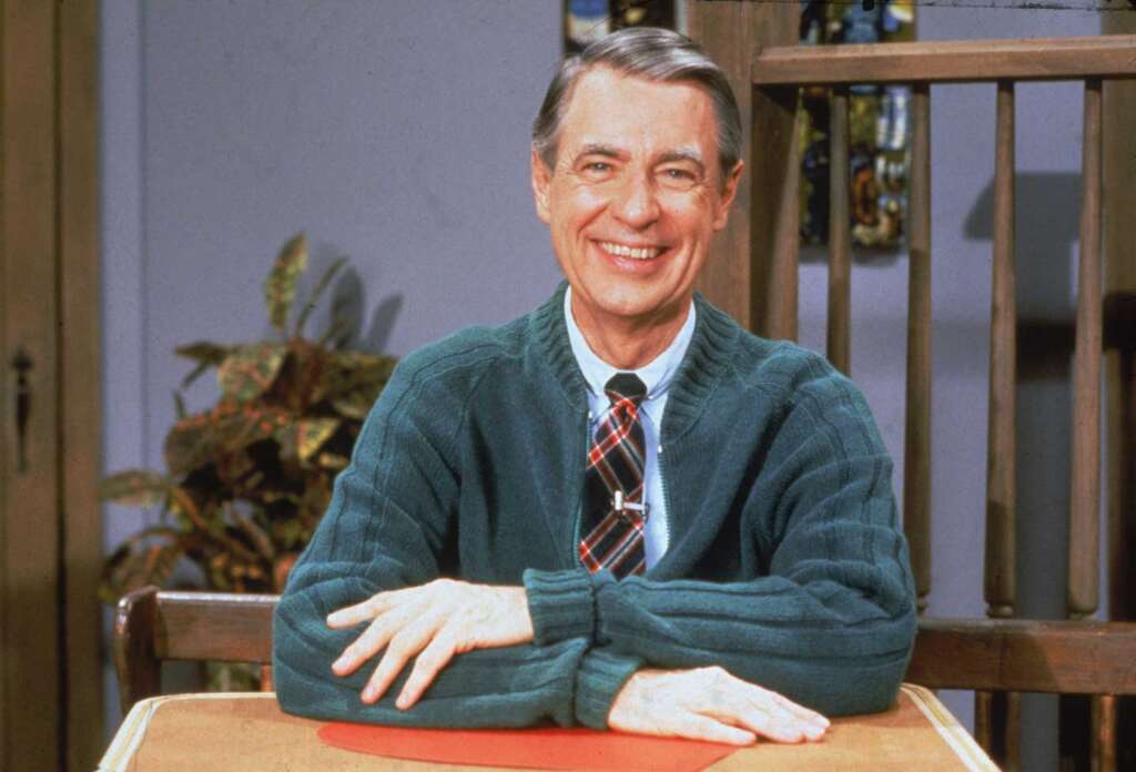 A smiling man with gray hair sits at a table, wearing a green cardigan and a plaid tie. There is a plant in the background and wooden slats beside him.