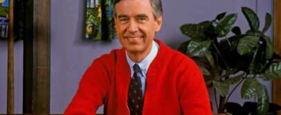 A smiling man with graying hair sits on a bench wearing a red cardigan over a tie and shirt. He is in a cozy room with a potted plant in the background and colorful curtains on the window.