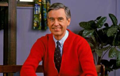 A smiling man with graying hair sits on a bench wearing a red cardigan over a tie and shirt. He is in a cozy room with a potted plant in the background and colorful curtains on the window.