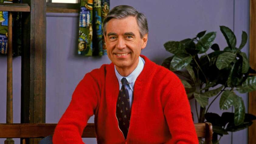 A smiling man with graying hair sits on a bench wearing a red cardigan over a tie and shirt. He is in a cozy room with a potted plant in the background and colorful curtains on the window.