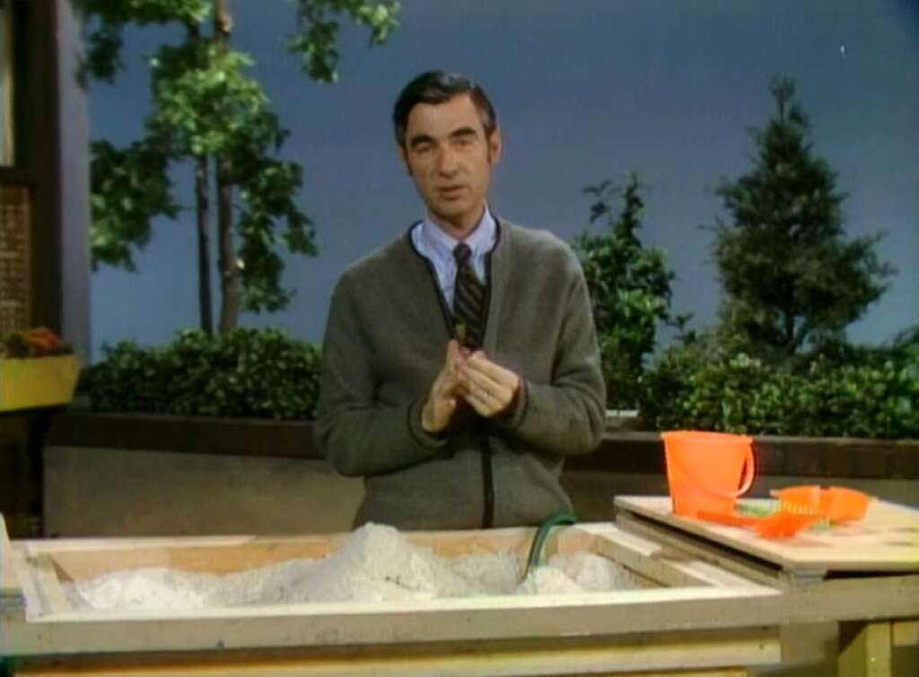 A man in a cardigan stands beside a sandbox with sand and a toy bucket. The setting appears to be outdoors with trees and greenery in the background. He is looking at the camera, gesturing with his hands.