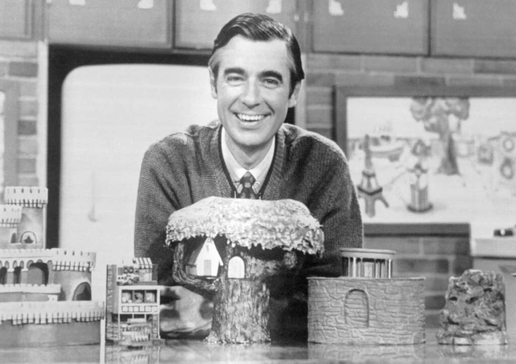 A person smiling broadly, wearing a sweater and tie, sits behind a table with various miniature buildings, including a treehouse. Shelves and a drawing are visible in the background, suggesting a creative or educational setting.