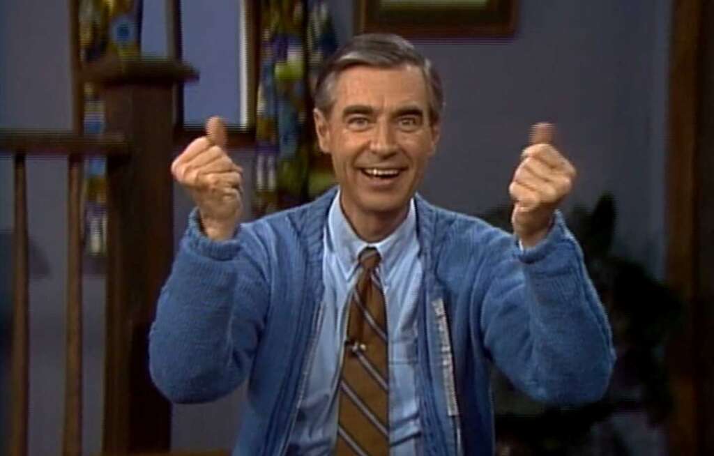 A man with gray hair, wearing a blue cardigan, shirt, and striped tie, smiles and gestures with both hands. He is seated indoors, with a staircase and patterned curtain behind him.