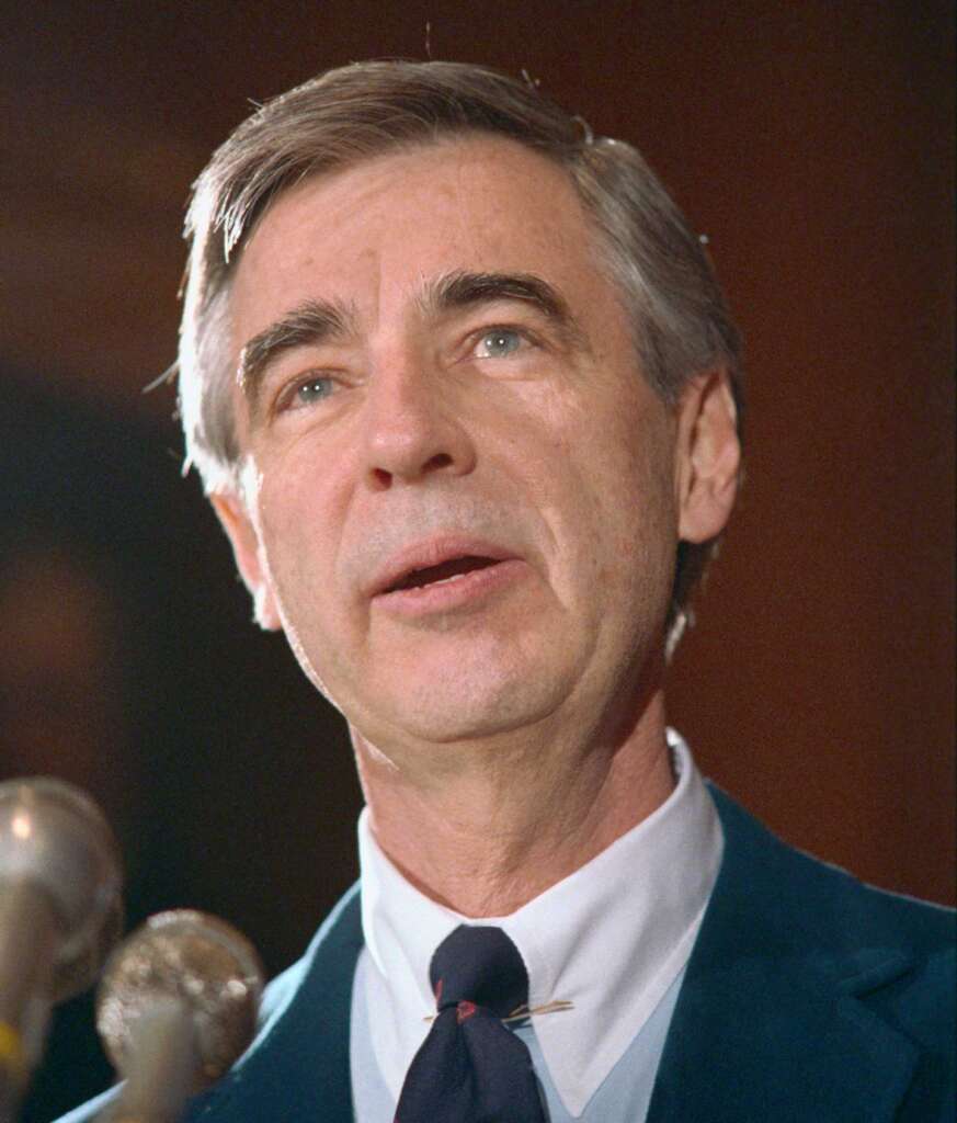 A middle-aged man with gray hair is speaking into several microphones, wearing a dark suit, white shirt, and tie. His expression is calm and focused, and the background is softly blurred.