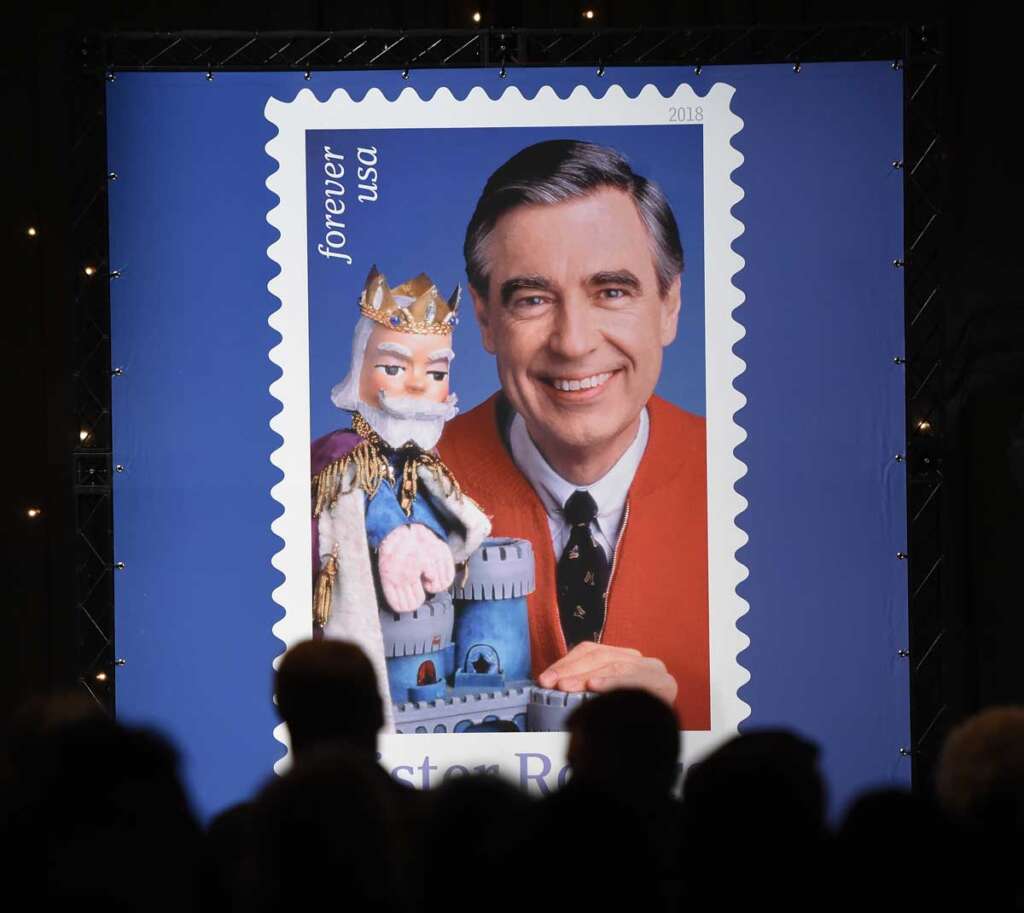 A large display of a postage stamp featuring a man in a red sweater and tie, smiling and posing with a puppet king. Silhouettes of people are visible in the foreground, facing the stamp.