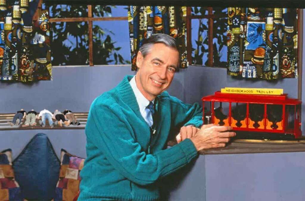 A man in a green sweater and blue tie smiles while holding a model of a red trolley labeled "Neighborhood Trolley." Behind him are colorful curtains and small figurines on a ledge.