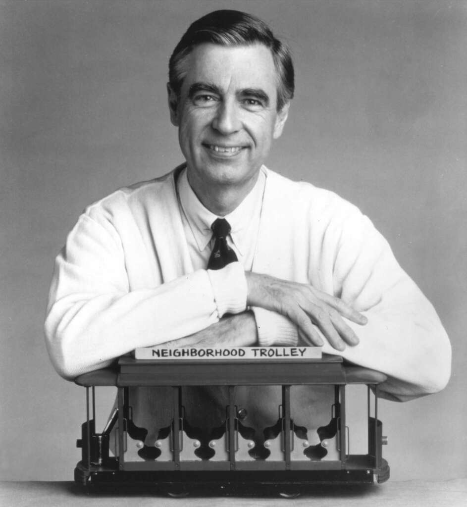 A black and white photo of a smiling person in a sweater and tie, arms resting on a model trolley labeled "Neighborhood Trolley." The person has a gentle expression and short, neatly combed hair.