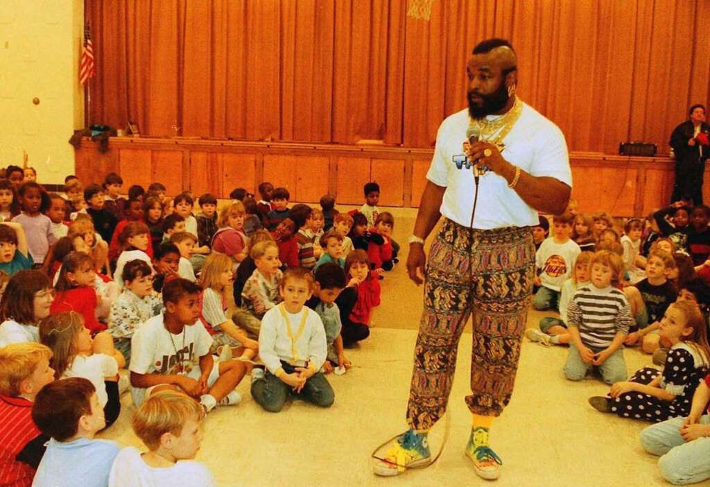 A man with a mohawk hairstyle, wearing a white T-shirt and patterned pants, stands with a microphone in a large room filled with seated children. Wood paneling and an American flag adorn the background.