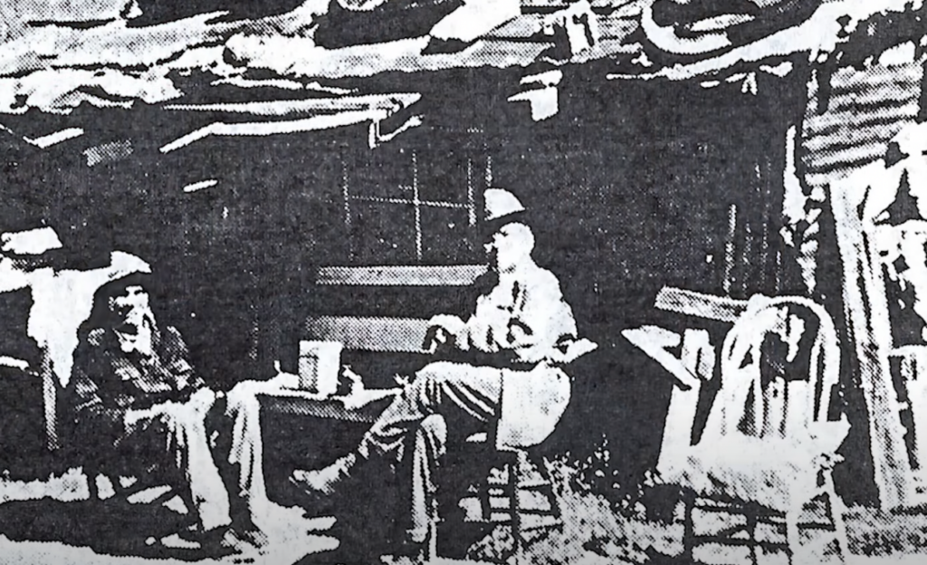 Black and white photo of two men sitting on chairs outside a rustic wooden shack. The man on the left leans back with a hat, while the man on the right wears a brighter hat and suit. Various objects and furniture surround the scene.