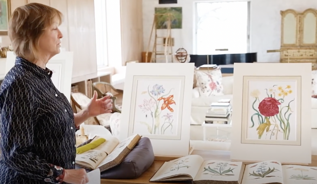 A person stands next to a table displaying botanical illustrations and open books. Two framed artworks of flowers, including lilies and peonies, are prominently featured. The setting is a well-lit room with a grand piano and floral-patterned furniture.