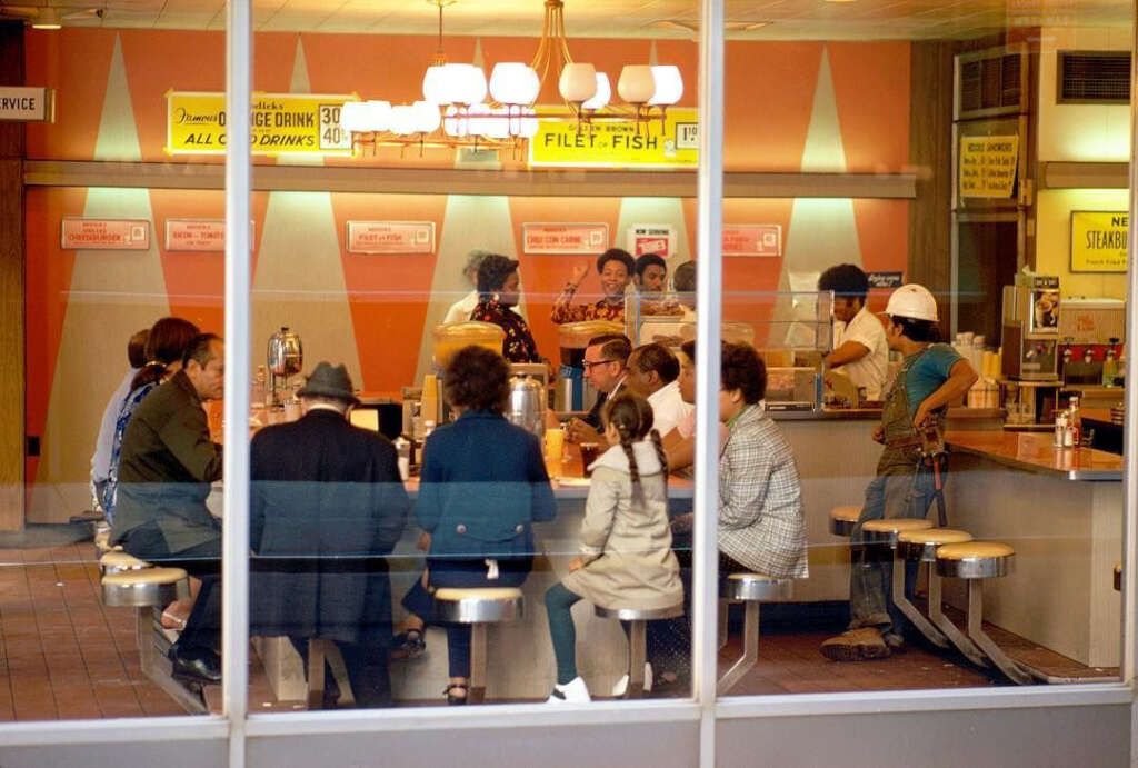 A retro diner scene with people seated at a counter and on stools, engaging in conversation. The interior is decorated in warm tones, and signs for drinks and food are visible. A soft overhead light illuminates the setting.