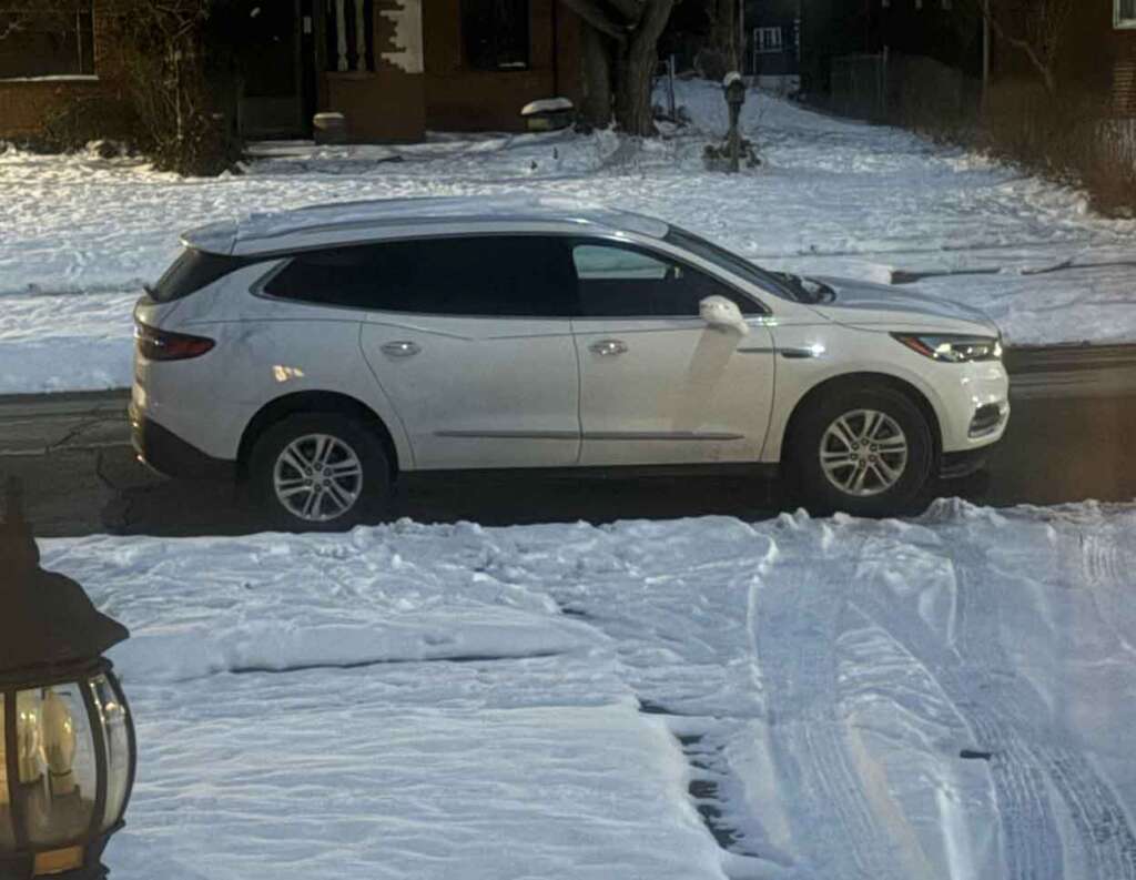 A white SUV is parked on a snow-covered street in a residential area. The ground is icy with tire tracks visible in the snow. A house and trees are in the background, and a decorative lamppost is in the foreground.