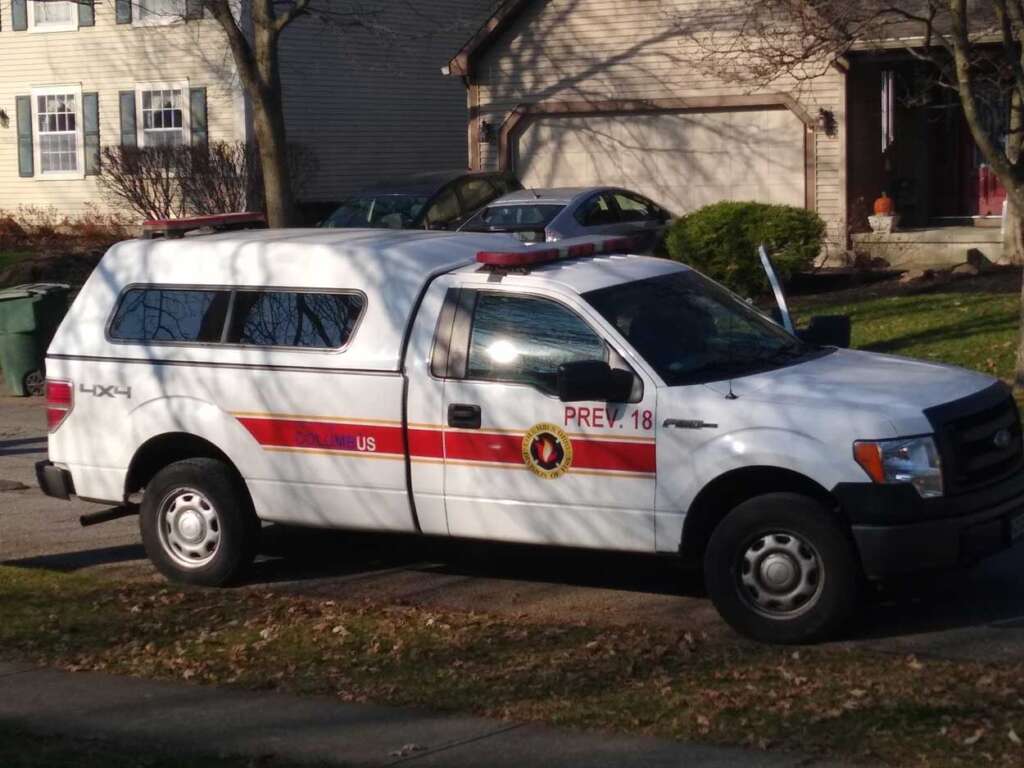 A white fire prevention truck is parked on a residential street. The truck has a red stripe, a light bar on top, and "Prevention" decals. Houses and trees are in the background.