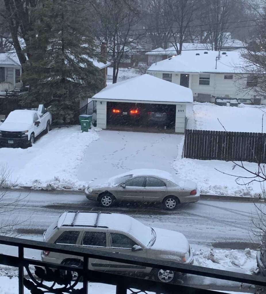 Snowy suburban scene with a house and a garage. Cars are on the driveway and street, all dusted with snow. Footprints are visible leading from the garage to the street. Trees and houses are in the background under overcast skies.