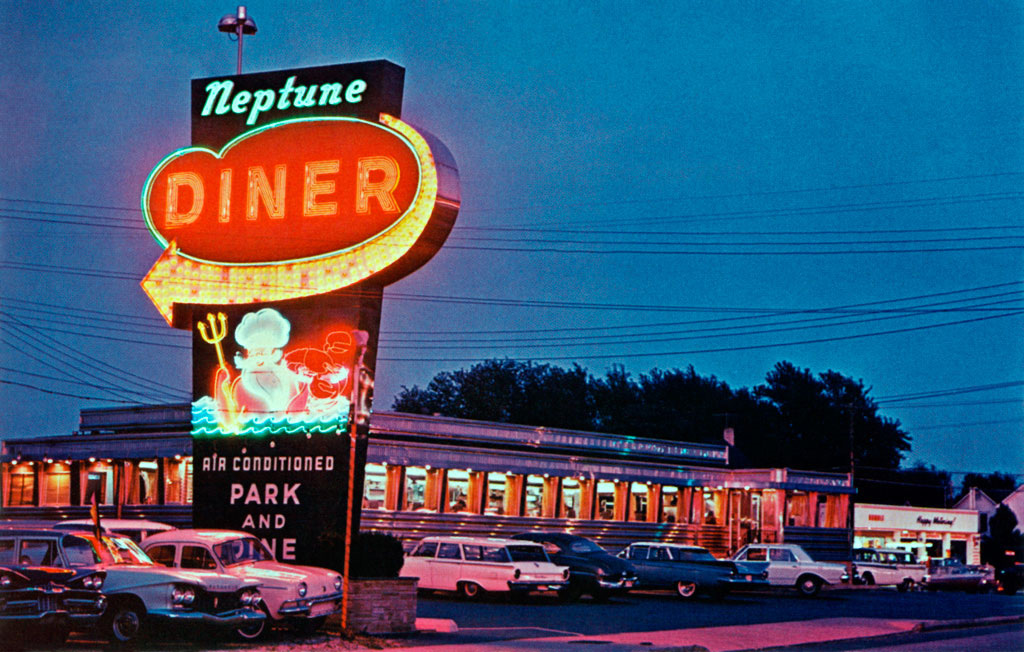 Vintage diner with a neon sign reading "Neptune Diner" featuring a trident-wielding character. Cars are parked outside along a well-lit street at dusk.