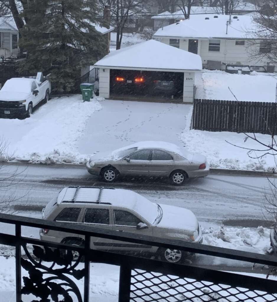 A snowy suburban scene with snow-covered cars parked along a street. A driveway leads to a garage with two red lights visible inside. Trees and houses are in the background, and snow covers the ground and rooftops.