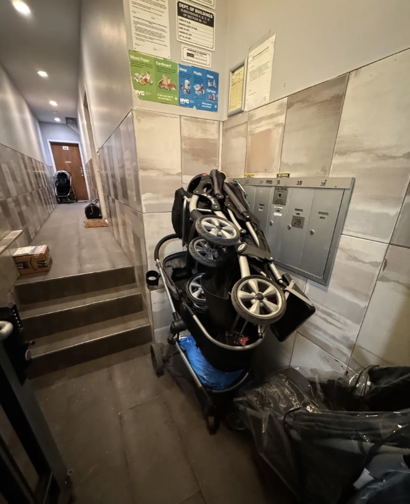 A narrow hallway with tiled walls, featuring stacked strollers near a set of metallic mailboxes. Notices are posted on the wall above the strollers. A small staircase is visible in the background.