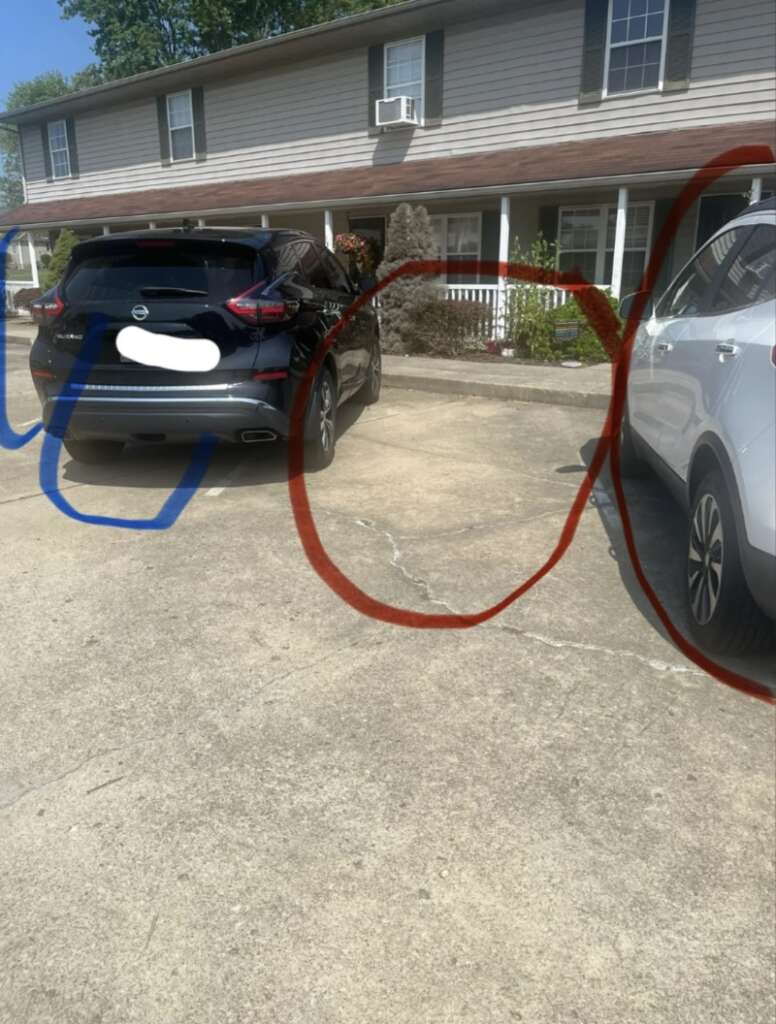 Two cars parked in a driveway outside a multi-story residential building. The left car is black, and the right car is white. Red and blue scribbles partially cover the image, with some sections of car license plates obscured.