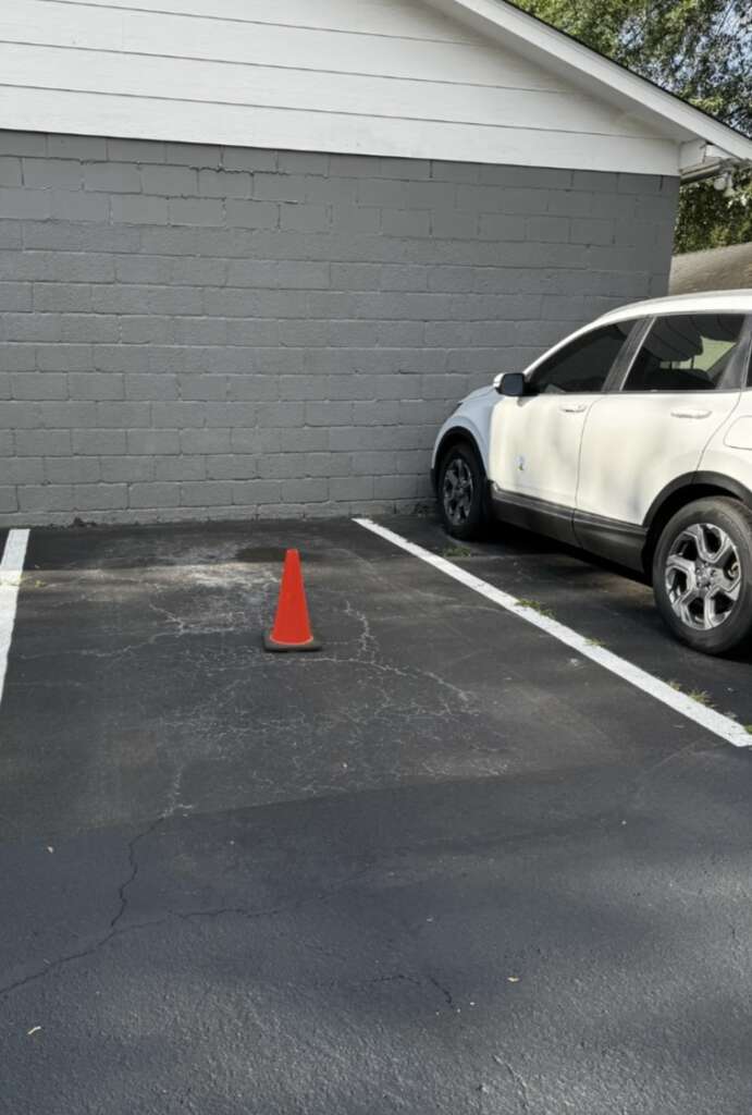 An empty parking space with a single orange safety cone placed in the middle. A white SUV is parked in the adjacent space next to a gray brick wall.