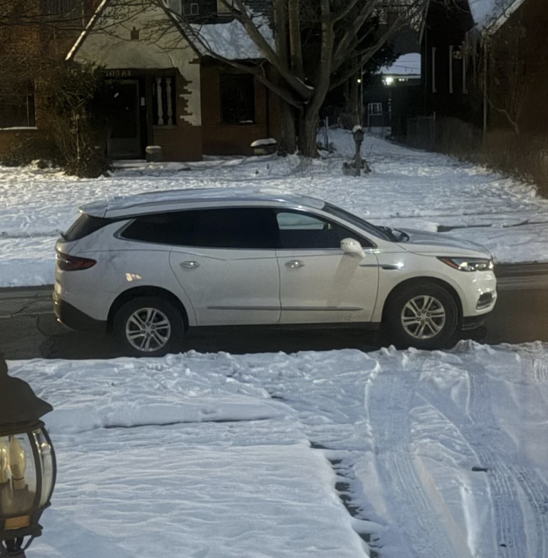 A white SUV is parked on a snowy street in front of a house with a porch. Snow covers the ground and surrounding yards. It's evening or early morning as the streetlights are on. A decorative lamp post is partially visible in the foreground.