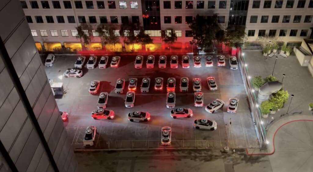 A rooftop view of a parking lot at night, where multiple white cars are arranged to spell out the word "TAXI". The surrounding buildings are illuminated, and red brake lights are visible on several cars.