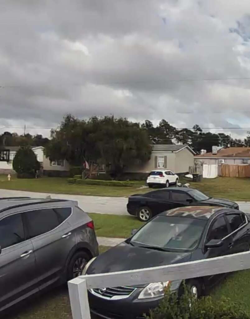 A suburban street with a few parked cars in a driveway and on the road. A gray SUV and a black sedan are in the foreground. Houses with lawns and trees are in the background under a cloudy sky.