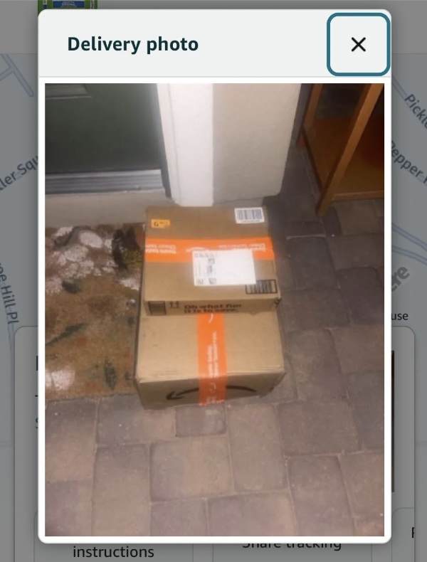 Two cardboard packages are stacked on a doorstep. One box has an orange stripe and a shipping label. The packages are placed on a patterned doormat in front of a green door, on a tiled surface.