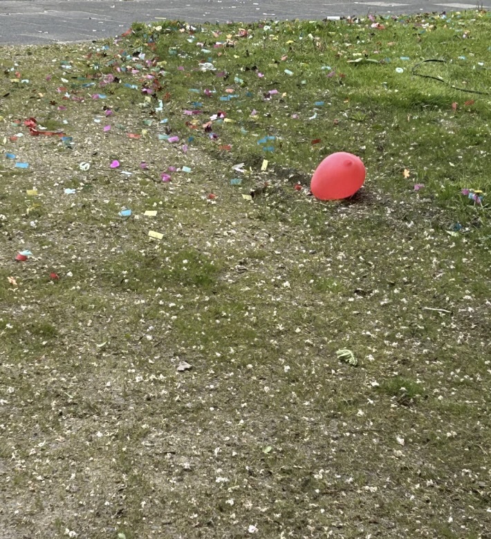 A red balloon lies on a grassy and gravelly area, surrounded by colorful small pieces of paper and confetti scattered across the ground. In the background, a paved surface is visible.