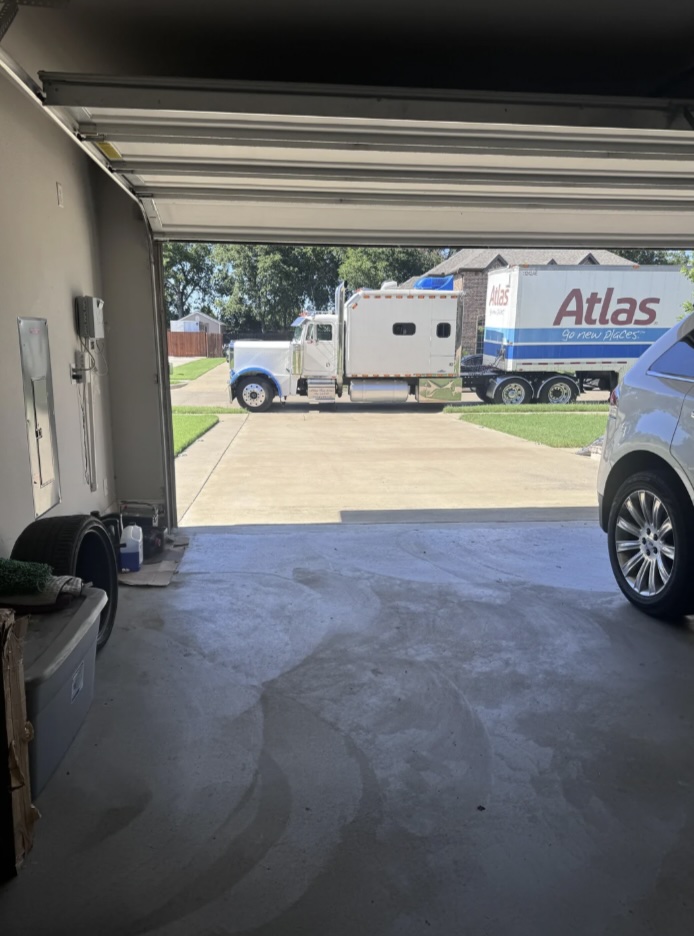 View from inside a garage looking out to a driveway where a large Atlas moving truck is parked. Part of a blue vehicle and various items are visible inside the garage. Trees and grass can be seen in the background.