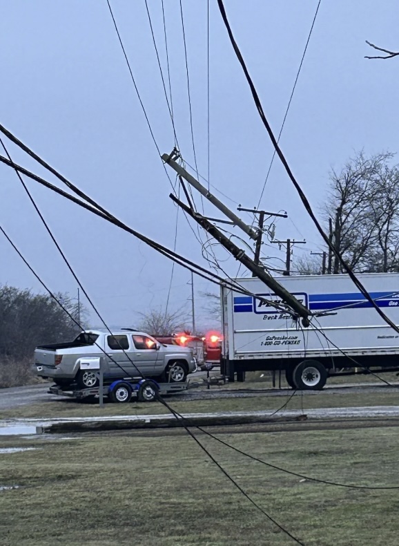 A utility truck is caught under fallen power lines, with a broken pole resting on top. Police lights flash in the background on a gray, overcast day. Trees and grass surround the scene.