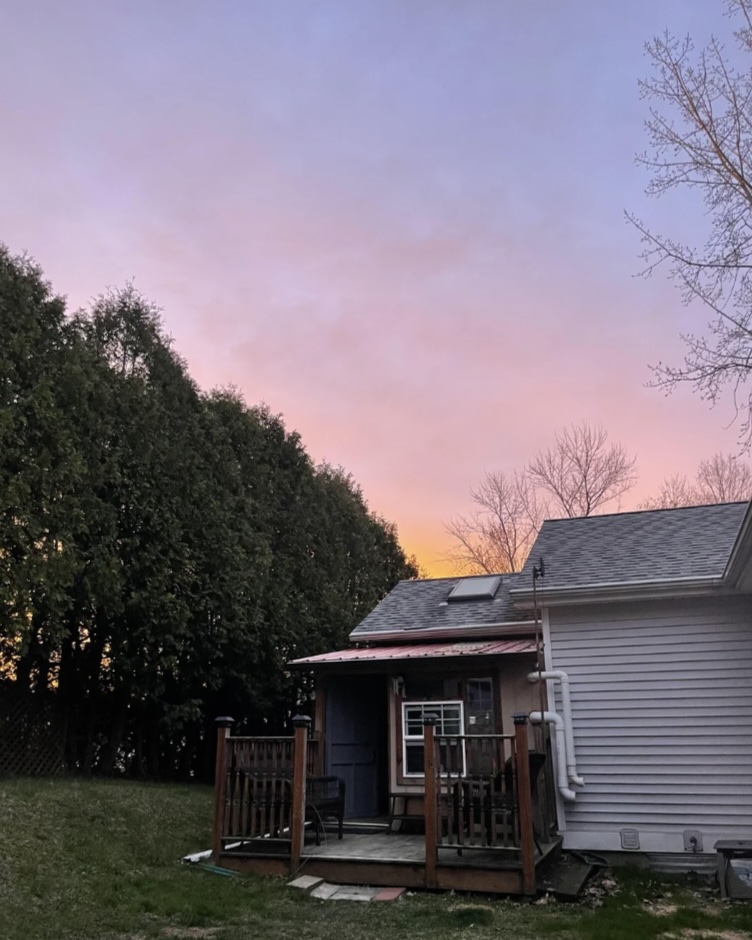 A cozy backyard scene at dusk features a small wooden deck attached to a house with a tree and hedge-lined background under a pink and purple sky.