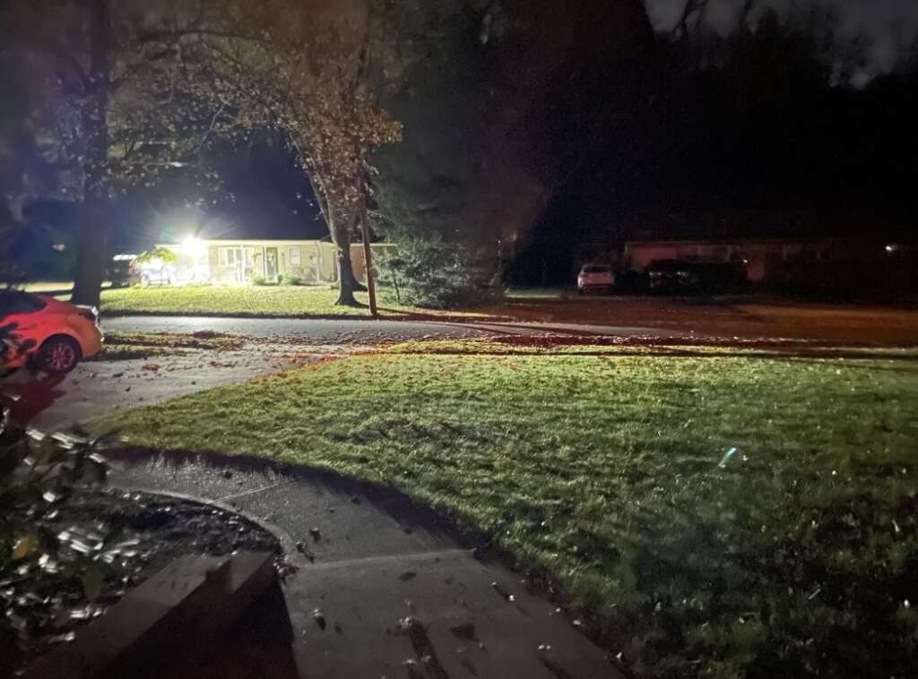 A dimly lit suburban street at night, with a grassy lawn and a sidewalk in the foreground. A house in the background is illuminated by bright exterior lights. A car is partially visible on the left, and trees are scattered throughout.