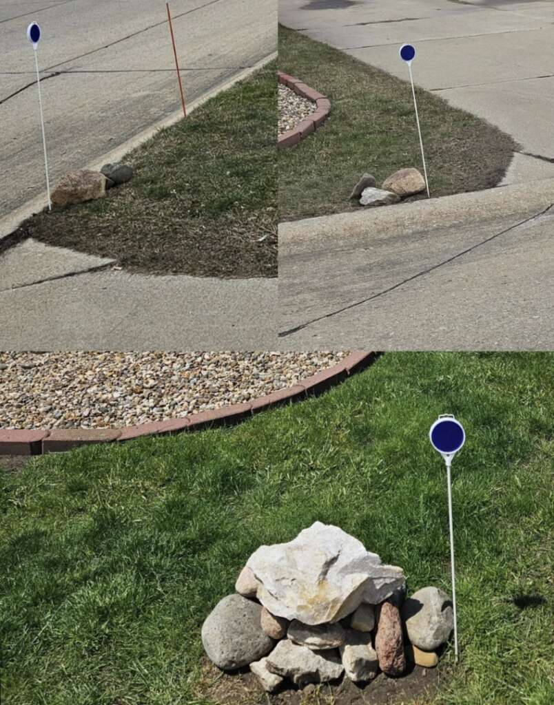 Three roadside markers with blue circles on white poles: one in a patch of dirt, one beside a driveway entrance, and one surrounded by rocks on grass. Each marker is next to a stone or small rock pile.